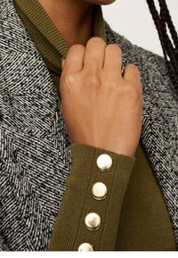 Blazer de pata de gallo gris con un suéter de cuello alto acanalado verde. El suéter presenta acentos de botones dorados en el puño de la manga.