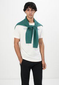 Junger Mann mit schwarzen Locken, der ein weißes Grafik-T-Shirt, schwarze Hosen und einen türkisfarbenen Pullover um die Schultern trägt, steht vor einem weißen Hintergrund.