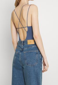 Niebieski bodysuit o prążkowanej strukturze z cienkimi ramiączkami i krzyżowym designem na plecach, w połączeniu z szortami dżinsowymi z wysokim stanem w odcieniu spranego denimu, z skórzaną naszywką z tyłu.