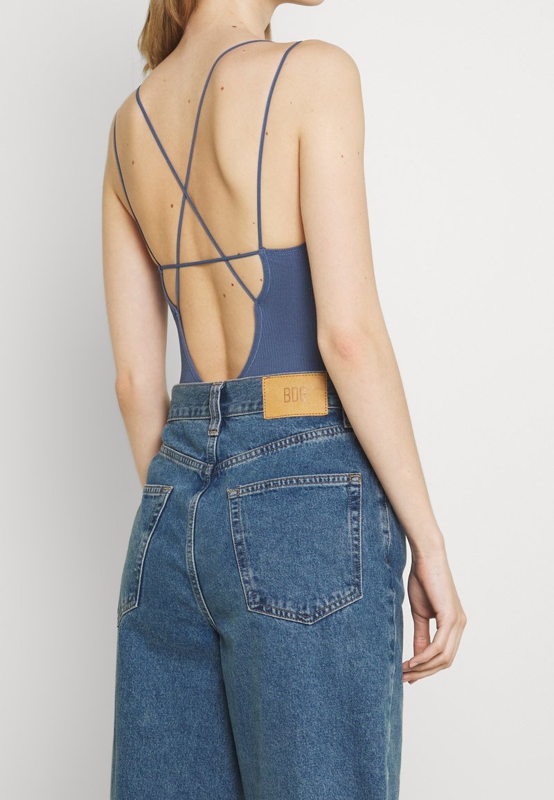 Niebieski bodysuit o prążkowanej strukturze z cienkimi ramiączkami i krzyżowym designem na plecach, w połączeniu z szortami dżinsowymi z wysokim stanem w odcieniu spranego denimu, z skórzaną naszywką z tyłu.