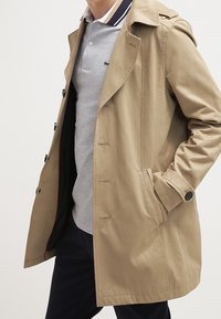 Beżowy trencz wykonany z fakturowanej tkaniny, z podwójnym zapięciem z przodu, wyciętym kołnierzem i bocznymi kieszeniami. Nosi się go na szarym polo.