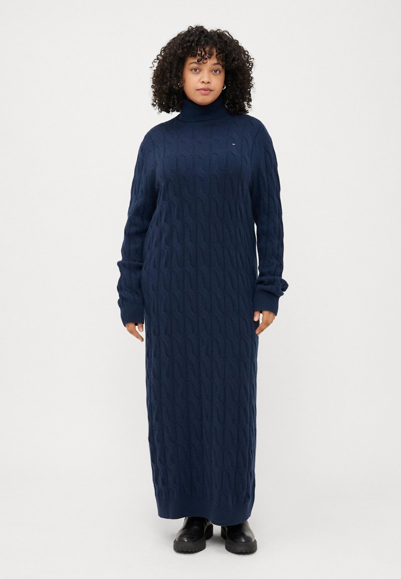 Frau mit lockigem Haar, die ein langes, navyblaues, strukturiertes Kleid und schwarze Stiefel trägt, steht vor einem schlichten weißen Hintergrund.