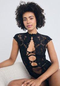 Black lace bodysuit featuring floral patterns, high neckline, and cut-out details with gold hardware accents. Sheer texture with cap sleeves.