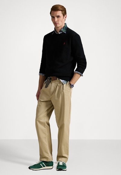 Suéter preto, camisa xadrez, calças bege, ténis verdes com detalhes brancos. Modelo está de pé com as mãos nos quadris, fundo neutro.