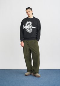 Črn pulover s belim grafičnim dizajnom, v kombinaciji z olivno zelenimi širokimi hlačami in bež natikači, na modrem preprogem tleh.