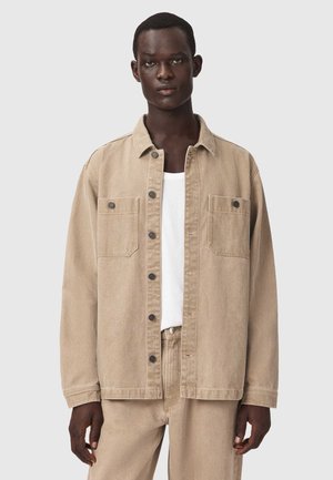 Modèle masculin portant une veste en jean beige avec des boutons noirs et un pantalon beige assorti sur une chemise blanche, debout devant un fond uni.