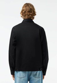 Melna ikdienas jaka ar gludu tekstūru, garām piedurknēm un apkakli; tajā ir pogas pie piedurkņu galiem un taisna apakšmalu.