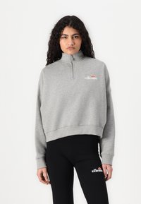 Felpa grigia corta con colletto alto, chiusura a zip e logo ricamato. Abbinata a leggins neri con dettaglio logo laterale.