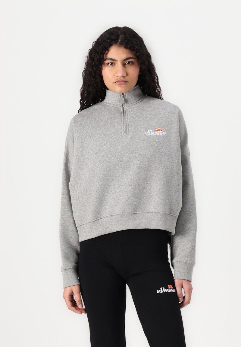 Sudadera gris cortada con cuello alto, cierre de cremallera y logo bordado. Combinada con leggins negros que presentan un detalle del logo en el lateral.