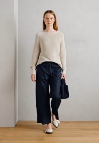 Beige hřejivý pletený svetr, volné námořnické kalhoty, krémové baleríny s mašlí a černá strukturovaná kabelka, stojící na dřevěné podlaze.