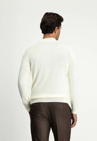 Helles cremefarbenes Pullover mit Rundhalsausschnitt, langen Ärmeln und gerippten Bündchen, kombiniert mit braunen Hosen mit zwei Knöpfen auf der Rückseite.