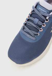 Kratki prikaz tamnoplave Skechers tenisice s gornjim dijelom od pletene tkanine, vezanjem na vezice i bijelim obloženim potplatom na bijeloj pozadini.