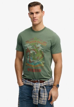 Uomo che indossa una maglietta verde del marchio Dragon con una grafica di drago e testo giapponese, jeans blu, camicia a quadri annodata in vita e orologio da polso.