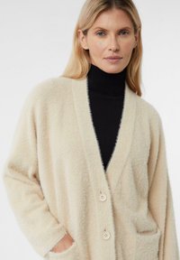 Beżowy sweter na drutach o miękkiej fakturze, z dekoltem w serek, dwoma kieszeniami z przodu i dwoma guzikami. Noszony na czarnym golfie.