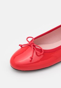 Ballerine en cuir verni rouge avec bout arrondi, nœud décoratif et bordure en tissu sur fond blanc.