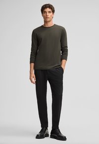Langsleeve, donkergroene shirt gecombineerd met zwarte, taps toelopende broeken en zwarte veterschoenen. Soepele stof met een relaxte pasvorm.