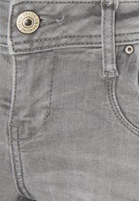 Primer plano de unos pantalones vaqueros de mezclilla gris que muestra el bolsillo delantero, el lazo del cinturón y el botón de metal grabado con "Genuine Denim Goods Lib."
