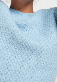 Pull en tricot bleu clair avec un motif tissé texturé. L'encolure ronde est bordée d'un bord côtelé, ce qui ajoute à sa douceur générale.