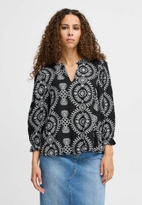 Blusa nera con motivi circolari bianchi e dettagli ricamati, caratterizzata da una scollatura a V e maniche a sbuffo, indossata sopra una gonna di jeans.