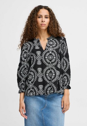 Schwarze Bluse mit weißen Kreis-Mustern und bestickten Details, mit einem V-Ausschnitt und Puffärmel, getragen über einem Jeansrock.