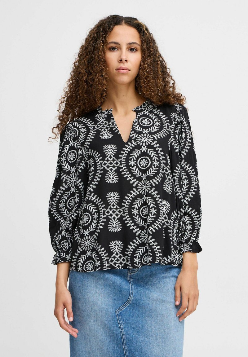 Blusa nera con motivi circolari bianchi e dettagli ricamati, caratterizzata da una scollatura a V e maniche a sbuffo, indossata sopra una gonna di jeans.