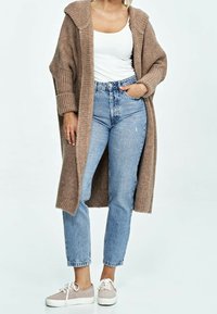 Long, beige knitted cardigan with wide sleeves, worn over a white tank top, paired with high-waisted light blue jeans and beige sneakers.