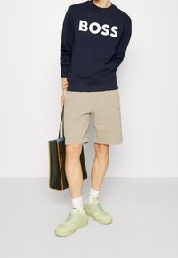 Námořnická mikina s bílým nápisem "BOSS", béžové šortky a zelené tenisky. Model drží tmavě zelenou tašku s oranžovými a modrými akcenty.