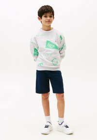 Grijze sweatshirt met groene grafische ontwerp en logo, gecombineerd met marineblauwe shorts en witte sneakers. Eenvoudige textuur en casual pasvorm.