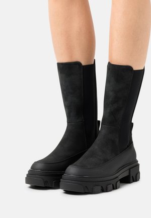 Bottes noires montantes en cuir suédé lisse, avec une semelle en caoutchouc épaisse et des panneaux latéraux élastiques pour un enfilage facile. Accents texturés sur le talon.