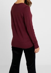 Maglione a maglia bordeaux con scollo rotondo e orlo a coste, abbinato a pantaloni neri con texture. Il tessuto sembra morbido e leggero.