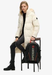 Zaino nero con dettagli rossi, logo sul davanti, materiale testurizzato; modello in giacca imbottita crema, maglione nero e jeans grigi.