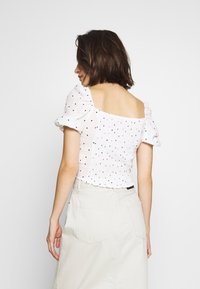 Kvinna med kort brunt hår klädd i en vit topp med prickar och puffärmar samt beige kjol, vänd bort från kameran mot en enkel ljus bakgrund.