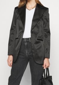 Blazer de satén negro con textura de satén, solapas con muesca, cierre de un solo botón y bolsillos laterales, combinado con jeans oscuros y un bolso negro.