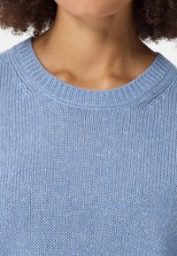 Hellblaues Strickoberteil mit strukturierter Oberfläche, das über einen gerippten Rundhalsausschnitt und weichen Stoff verfügt. Nahaufnahme, die den Ausschnitt und die Textur zeigt.