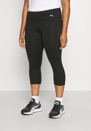 Person iført sorte Puma korte leggings, hvide sokker, sorte og hvide Puma sneakers og en hvid top, stående på en grå gulv.