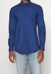 Maglietta a maniche lunghe di colore blu scuro con colletto a punta e chiusura con bottoni. Presenta bottoni neri e una trama liscia e uniforme.