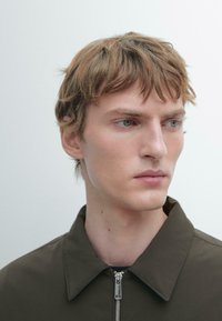 Ung mand med lysebrunt hår iført en mørkegrøn lynlåstrøje, der tænksomt kigger mod højre mod en ensfarvet lys baggrund.