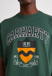 Zielona koszulka z bawełny z napisem "CARHARTT", logo w kształcie tarczy z pomarańczowymi i czarnymi akcentami oraz białą wstążką poniżej z napisem "THE LAZY DUCK."