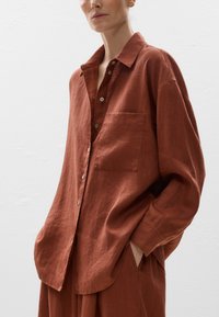 Camicia di lino color ruggine con vestibilità rilassata, design con bottoni, tasca sul petto e maniche arrotolate. Tessuto morbido e leggero lucido.
