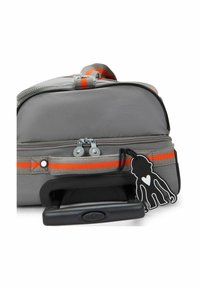 Bolsa de tela gris con rayas naranjas, cierre de doble cremallera y una etiqueta de mono de dibujos animados negra. Mango retráctil negro visible.