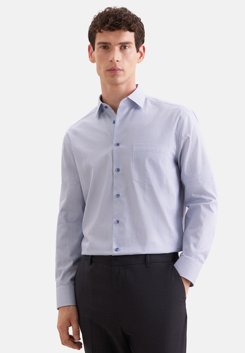 Homme portant une chemise à manches longues bleu clair à motifs, rentrée dans un pantalon noir, debout avec une main dans la poche.