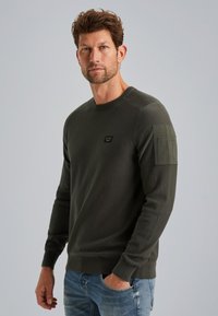 Maglione verde scuro a maniche lunghe, con scollo a girocollo e una tasca a toppa sulla spalla. Realizzato in un materiale a maglia testurizzato.