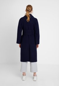 Langer marineblauer Mantel mit tailliertem Gürtel, weiten Ärmeln und einem strukturierten Design. Wird über hell karierten, gekürzten Hosen und weißen Sneakers getragen.