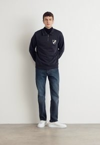 Pulover închiriu închis, cu guler cu fermoar, emblemă pe față, asortat cu blugi albaștri și adidași albi. Material neted, croială standard, design minimalist.