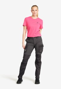 Kvinna som står med handen i fickan, klädd i en ljusröd kortärmad topp, mörkgrå cargobyxor och svarta skor, på en vit bakgrund.