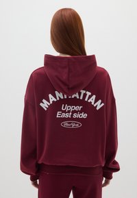 Felpa con cappuccio color bordeaux con testo stampato: "MANHATTAN Upper East Side New York" in bianco. Tessuto morbido, vestibilità rilassata.