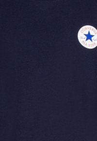 T-maica temno modre barve iz bombaža z okroglim izrezom in krožnim belim logotipom z modro zvezdo, rdečim besedilom ter gladko teksturo.