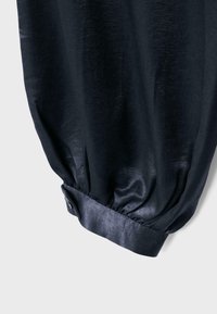 Sort skinnende langærmet trøje med en knaplukning ved manchet og samlet stof, der skaber en let puffet effekt nær håndleddet.