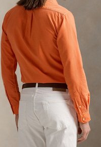 Orange skjorta med lång ärm och en veckad rygg, kombinerad med vita högmidjade byxor. Har knappmanschetter och ett brunt läderbälte.