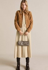 Giacca in suede marrone, abito plissettato beige, stivali marroni fino al ginocchio e borsa argentata. Texture morbide, silhouette classica e design semplice.